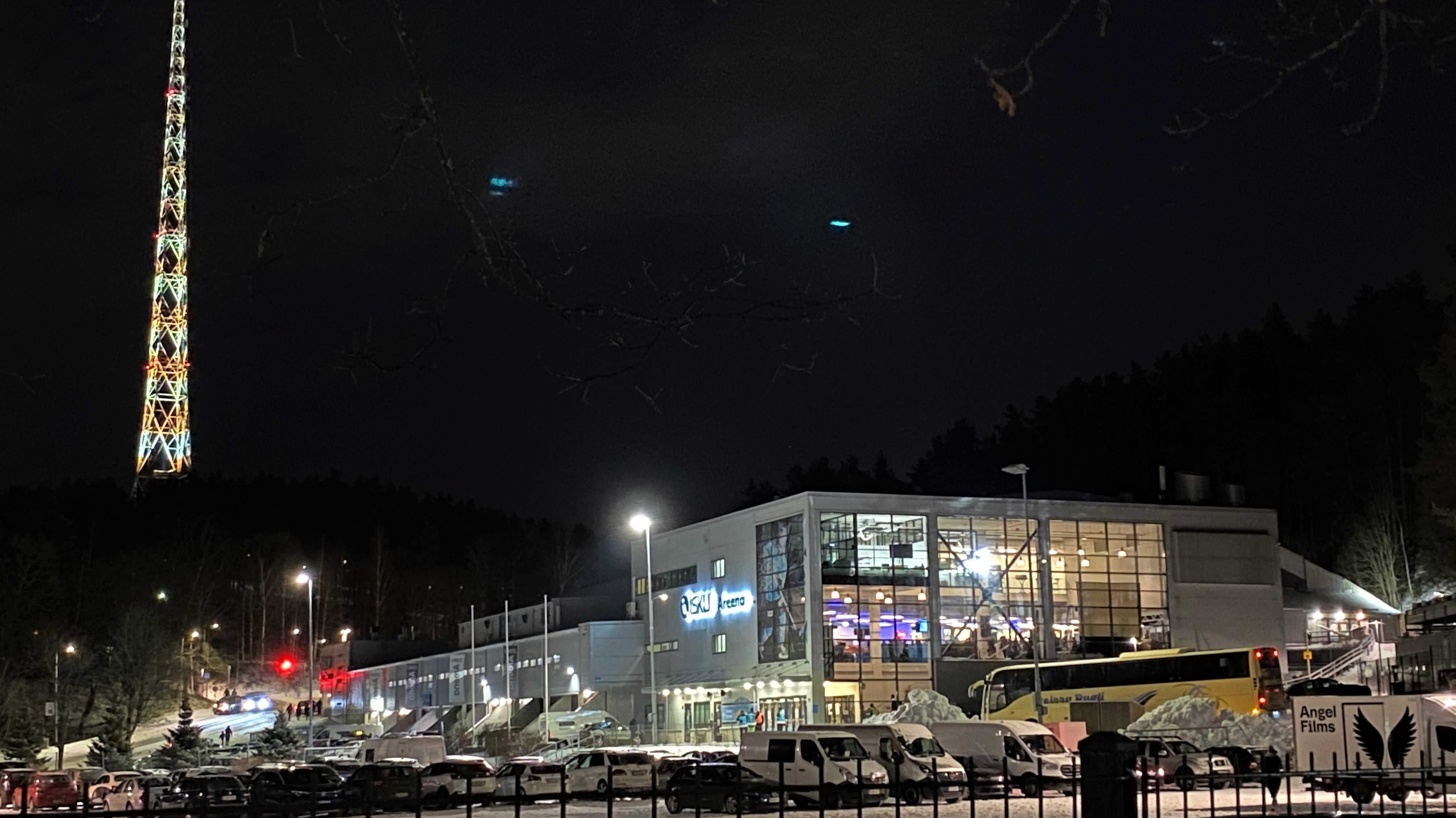 Iltakuva: pimeessä hohtaa Lahden radiomaston valot, kuvassa Lahden jäähalli jossa valot. Etualalla parkkipaikka täynnä autoja. 