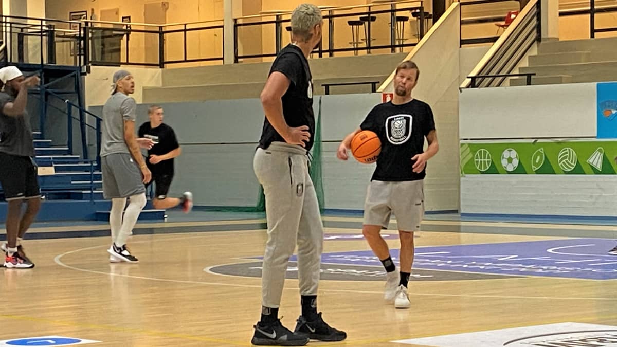 Mustapaitainen korisvalmentaja Pieti Poikola neuvoo harjoituksissa pelaajaa, valmentajalla kainalossaan koripallo. Pelaaja kuuntelee seisten kädet lanteillaan. 