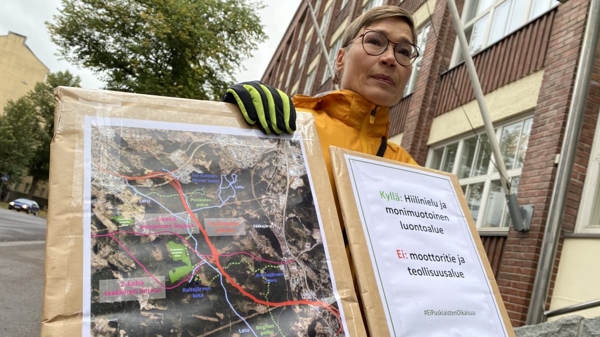 Kristiina Kaila pitelee kahta kylttiä kameran suuntaan. Toisessa kyltissä on moottoritien kartta, jonka alla teksti #eipuskiaisten oikaisua, ja toisessa teksti "Kyllä: Hiilinielu ja monimuotoinen luontoalue, EI: moottoritie ja teollisuusalue".