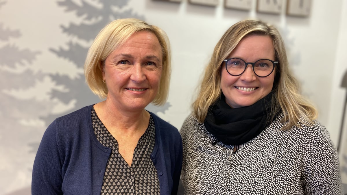 Lähikuvassa kaksi vaaleahkohiuksista naista, vasemmalla polkkatukkainen nainen sinisessä villatakissa ja oikealla hiukan nuorempi silmälasipäinen ja pitkätukkainen nainen. Molemmat hymyilevät. 
