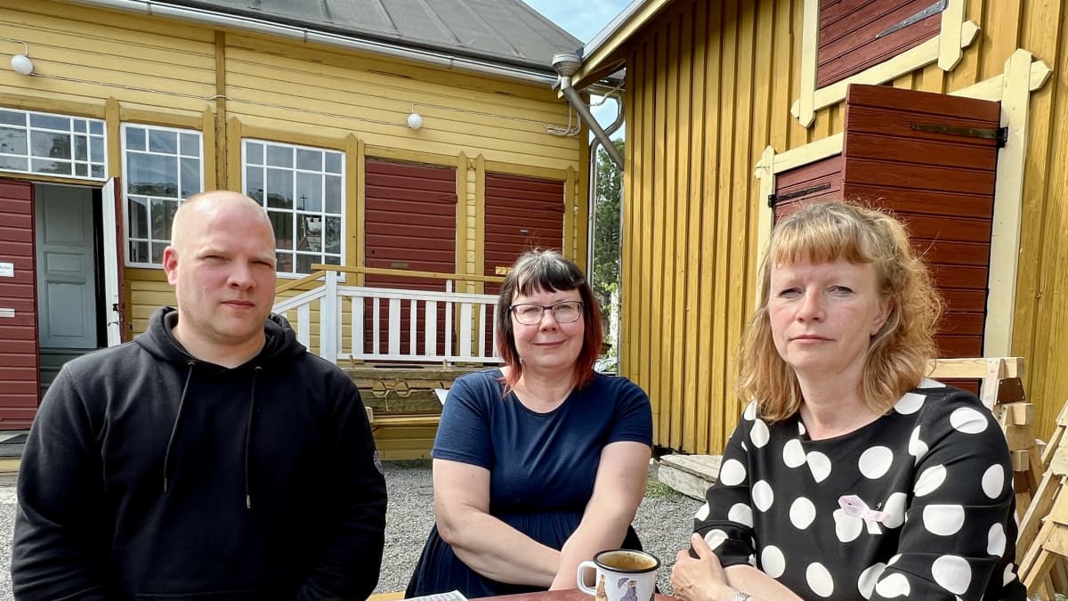 Antti Nyman, Heidi Huhtamella ja Katja Wallenius istuvat puisen terassipöydän ympärillä ja katsovat kohti kameraa. Taustalla on vanhoja puutaloja.