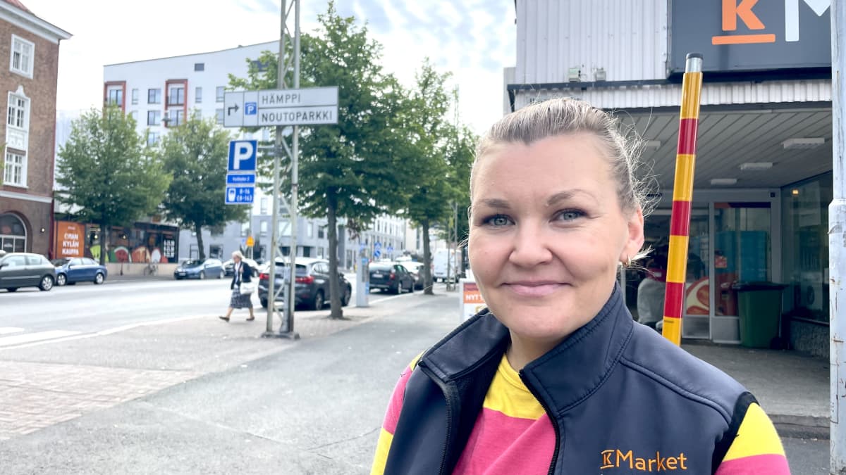 Nainen kelta-pinkissä paidassa ja kauppiaan harmaa liivi päällä seisoo K-Marketin edessä ja katsoo hymyillen kameraan.