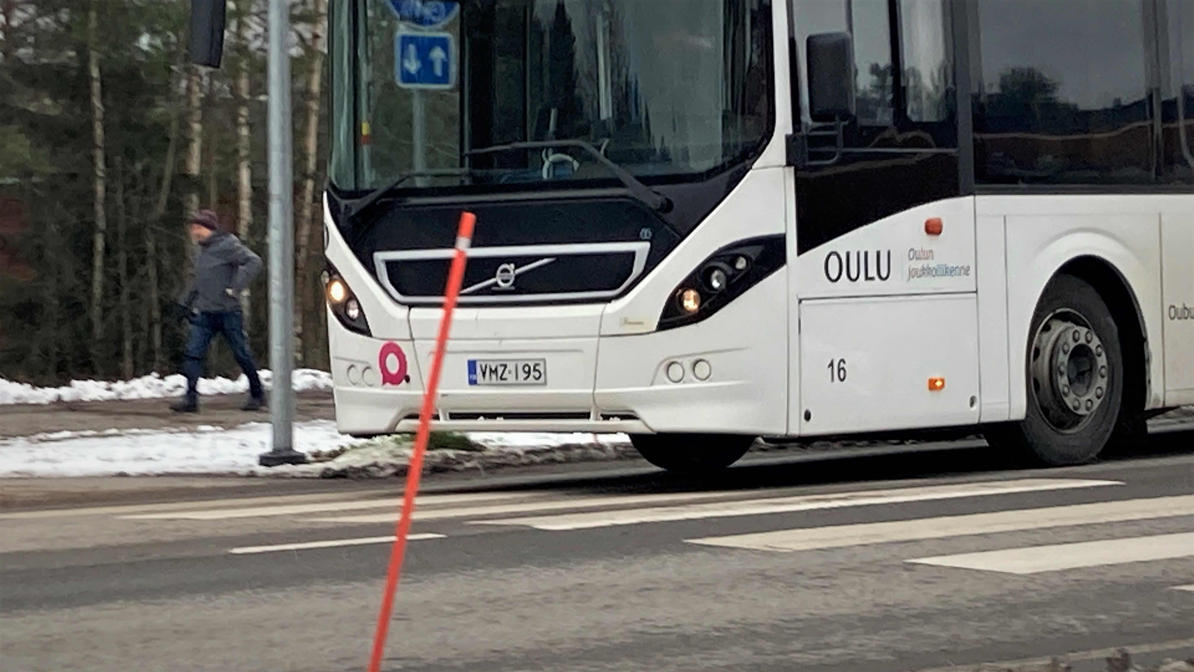 Oulun joukkoliikenteen valkoinen bussi ajaa tiellä. Taustalla jalankulkija kävelee jalkakäytävällä.