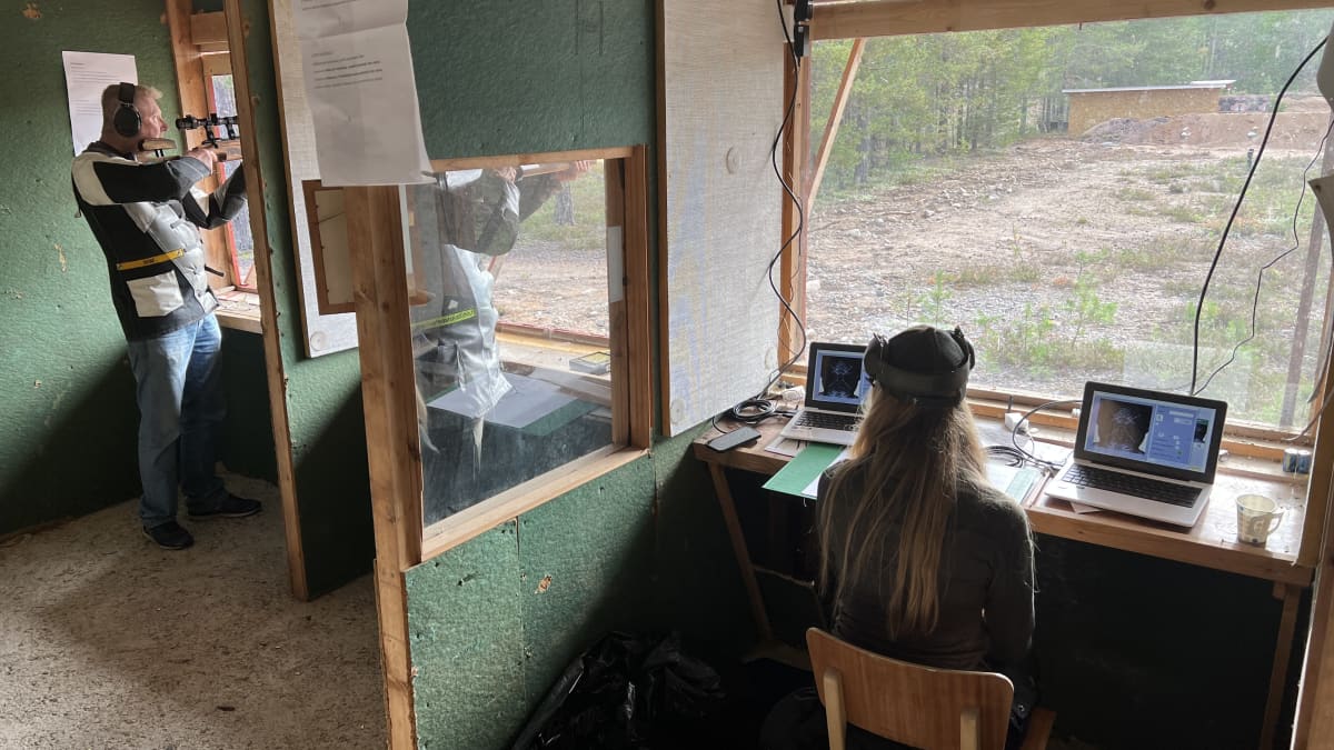 Kaksi miestä tähtää kivääreillä kohti maalitaulua ampumakopissa, jossa on isot ikkunat. Nainen tarkkailee monitoria.
