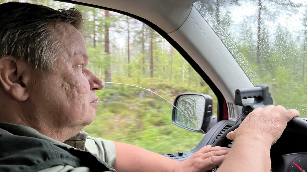 Harmaahiuksinen mies istuu autossa kädet ratissa. Puoliksi avoimesta ikkunasta näkyy metsää.