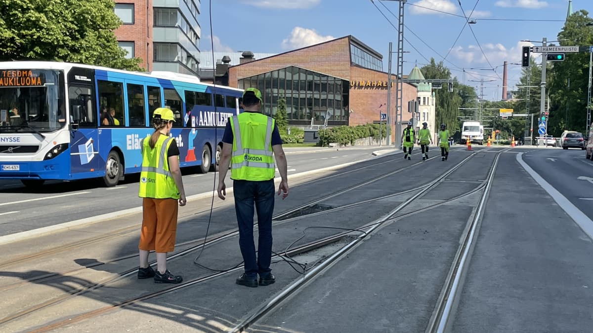 Huomioliiveihin ja kypäriin pukeutuneita ihmisiä seisomassa Tampereella ratikkakiskojen päällä. Keli on kesäinen ja aurinkoinen.