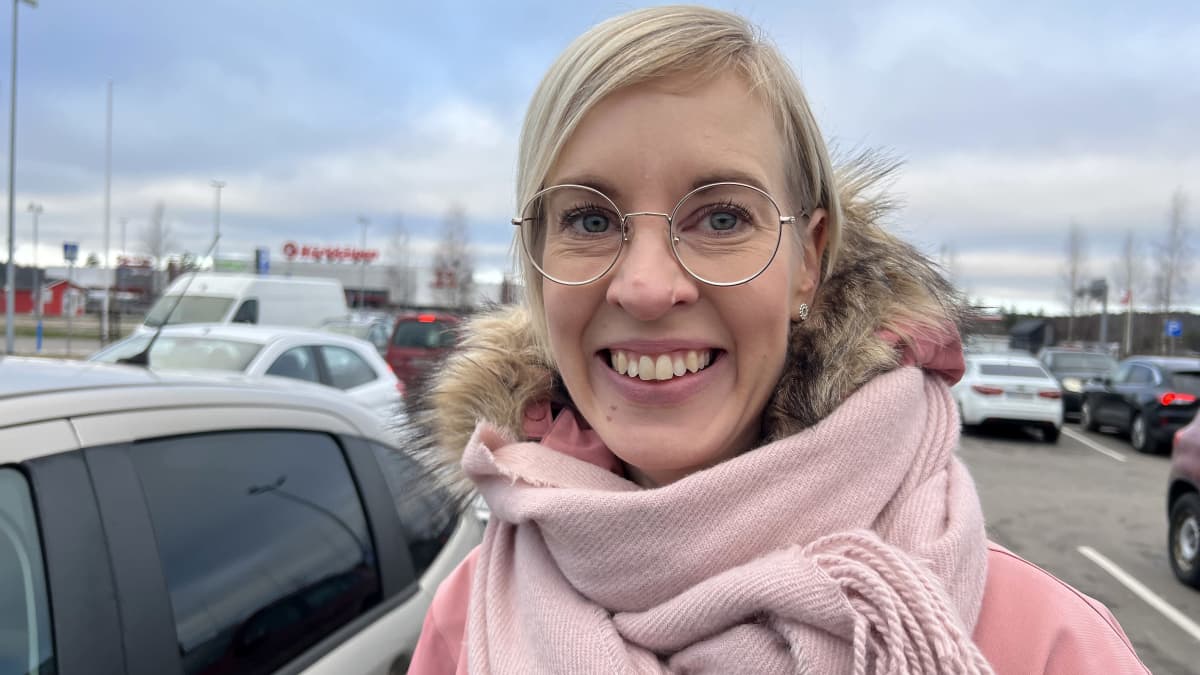 Lahtelainen Suvi Kivekäs kauppaostoksilla, kuva parkkipaikalla, Suvilla pyöreät isokokoiset silmälasit, vaaleat hiukset, hymyilee.
