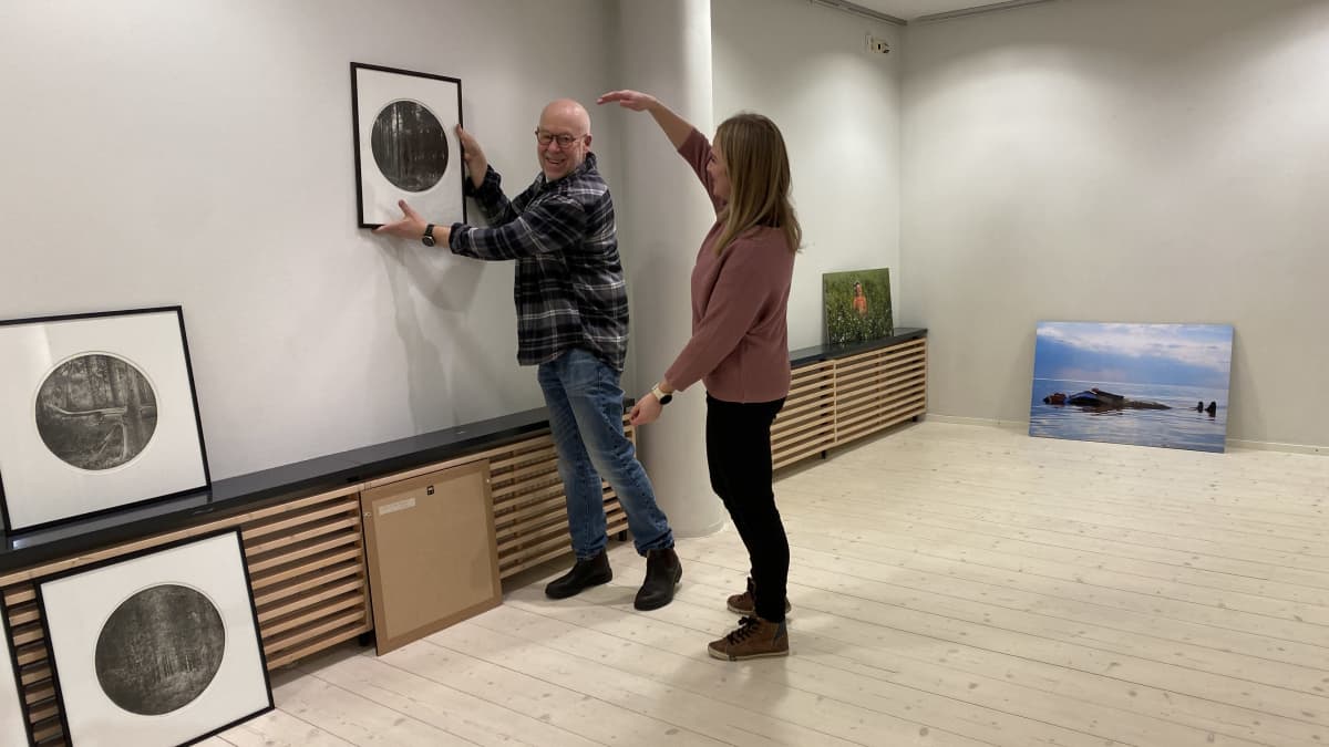 Vasemmalla Leif Strengell åitää työtään seinällä, oikealla Anni Laukka auttaa ripustuskorkeuden määrittelyssä.