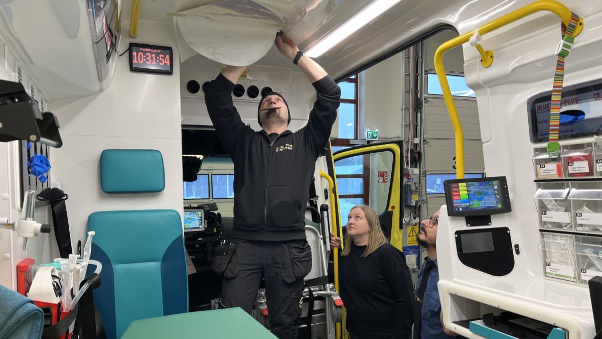 Olli Hyvönen teippaa tarraa ambulanssin kattoon, Heidi Anttonen ja Lasse Hakala seuraavat tilannetta ambulanssin ovelta.