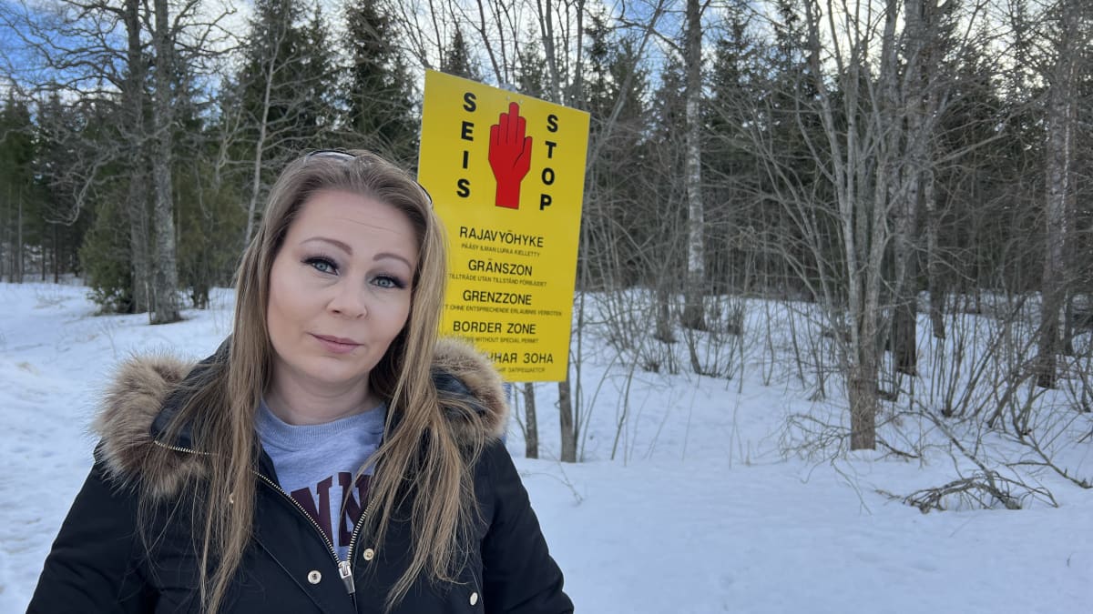 Nainen seisoo ulkona suuren kyltin edessä. Puna-keltaisessa kyltissä on punainen käsi ja siinä lukee Stop! Rajavyöhyke.