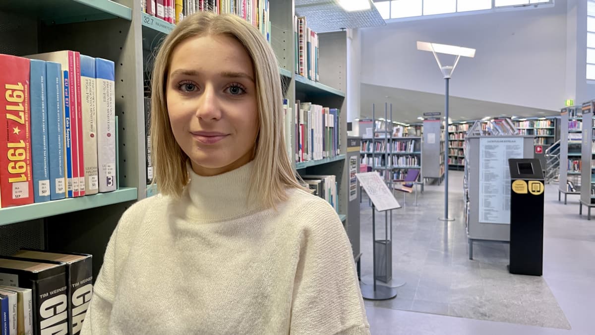 Vaaleahiuksinen nuori nainen seisoo kirjastossa kirjahyllyn vieressä ja katsoo kohti.
