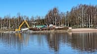 Kaivinkone tekee töitä veden äärellä kauniissa kevätsäässä. Taustalla näkyy puustoa ja työmaa-aitoja.