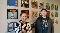 Näyttelyamanuenssi Elina Suutari ja näyttelyn kuratoinut levykauppayrittäjä Ari-Pekka Savolainen seisovat seinän edessä, jonne on ripustettu levynkansia. 
