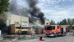 Teollisuusrakennus palaa mustaa savua tupruttaen. Rakennuksen edessä kadulla paloauto, jonka takaa suihkuaa vettä paloon.