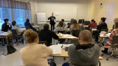 Luokka, jossa etualalla aikuiset suomenkielen opiskelijat istuvat selin, ja opettaja puhuu luokan edessä.