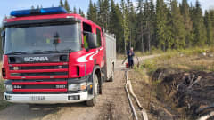 En brandman jobbar med en släckningsslang vid en brandbil. Bilen står parkerad på en skogsväg.