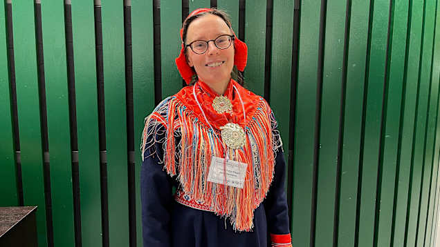 Nainen saamenpuvussa viherän seinän edessä.