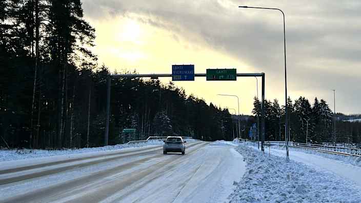 Auto ajaa tiellä, lunta on tiellä, tien vieressä on puita. 