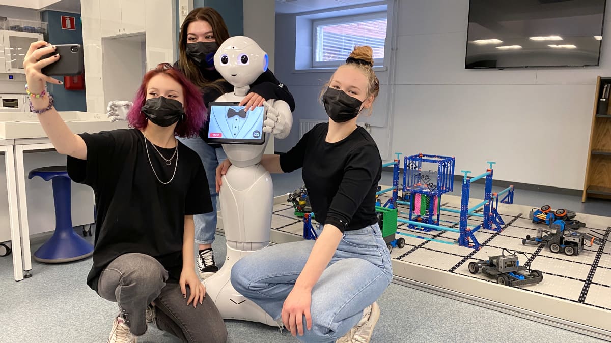 Jenna Hietakangas, Pinja Jokinen ja Sara Korkia-aho ottavat selfietä yhdessä ihmishahmoa muistuttavan humanoidirobotin kanssa.