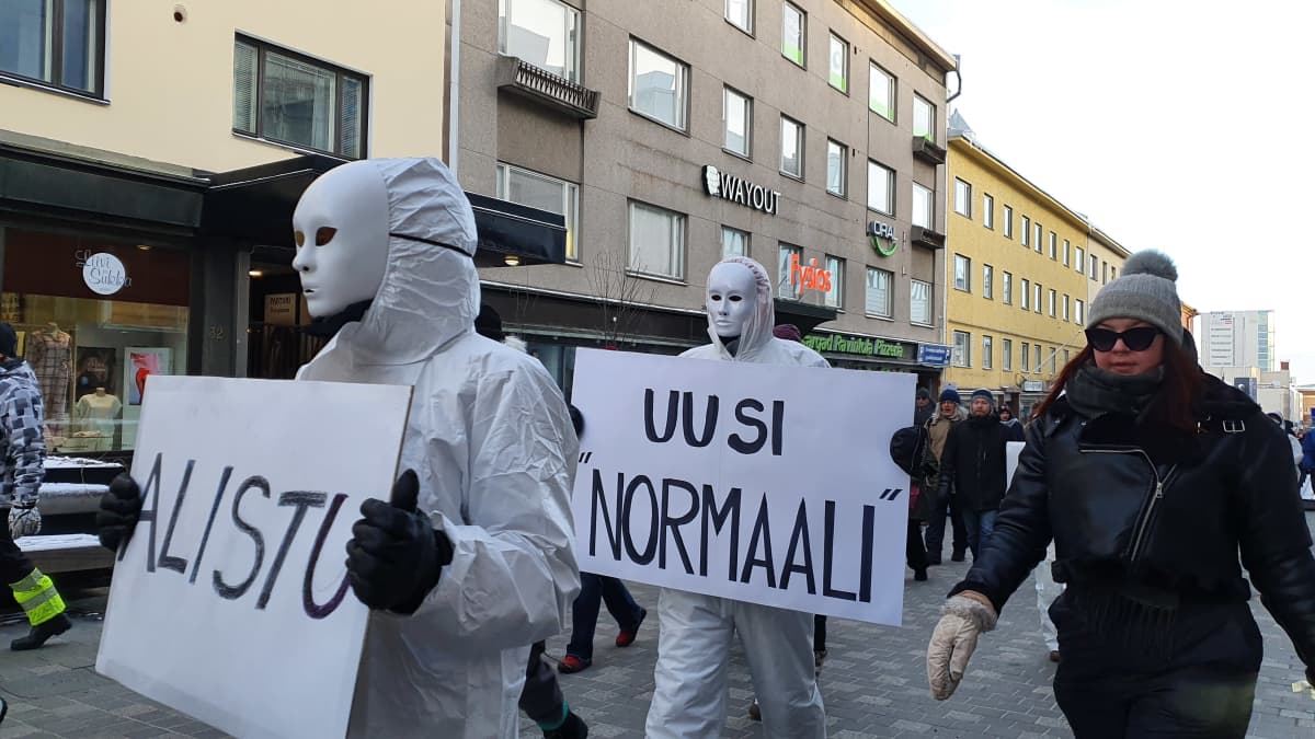 A person wearing white overalls holds a sign that reads "new normal".