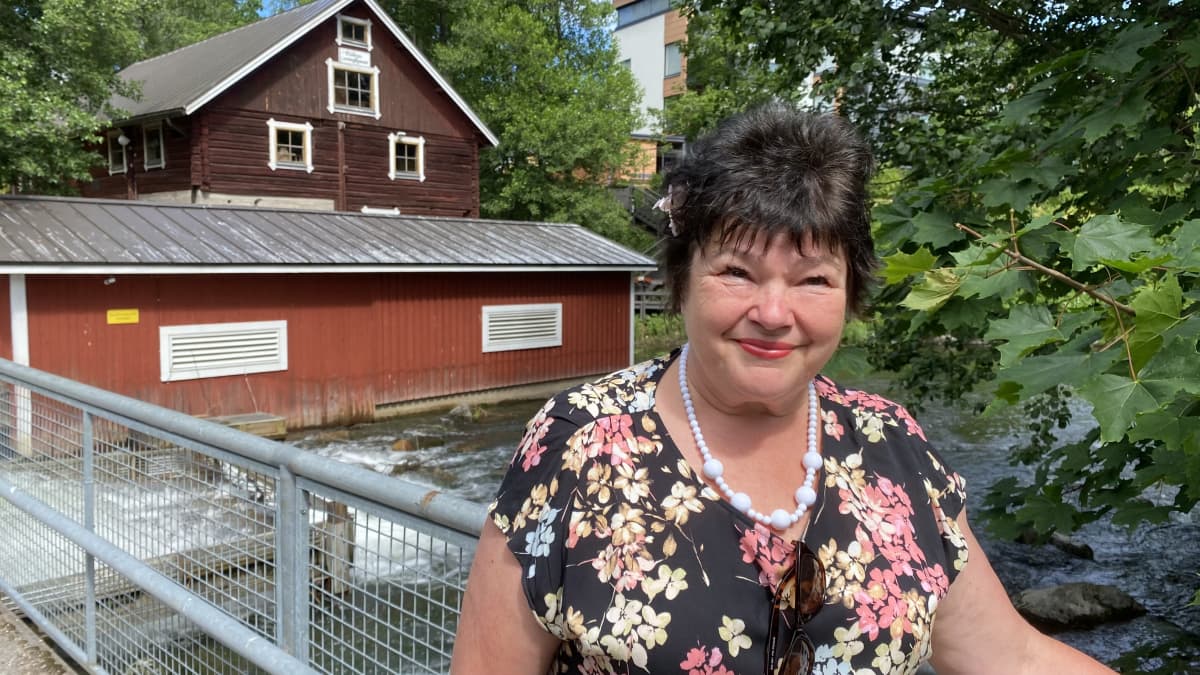 Kukkamekkoon pukeutunut nainen nojaa rautaiseen sillankaiteeseen, taustalla vanhoja puurakennuksia