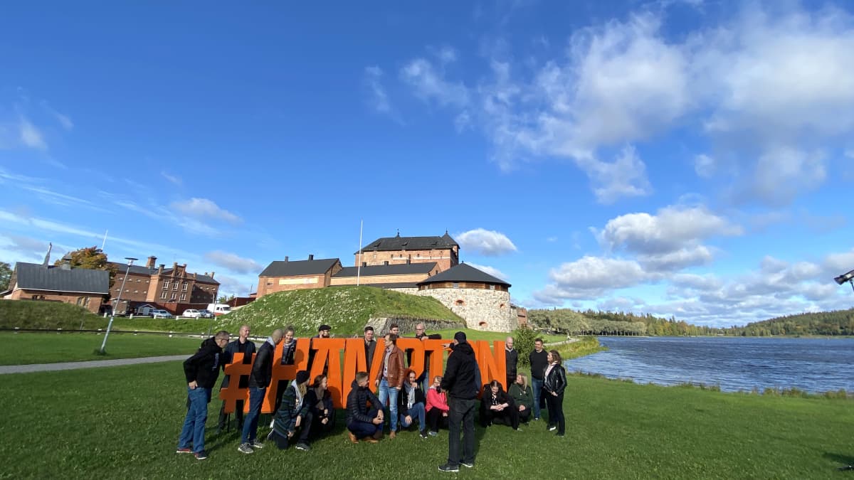 Joukko ihmisiä ryhmittäytyy oranssisten Hämpton-kirjaimien luo puistossa. Takana vanha linna, sinitaivas.ja järvi.