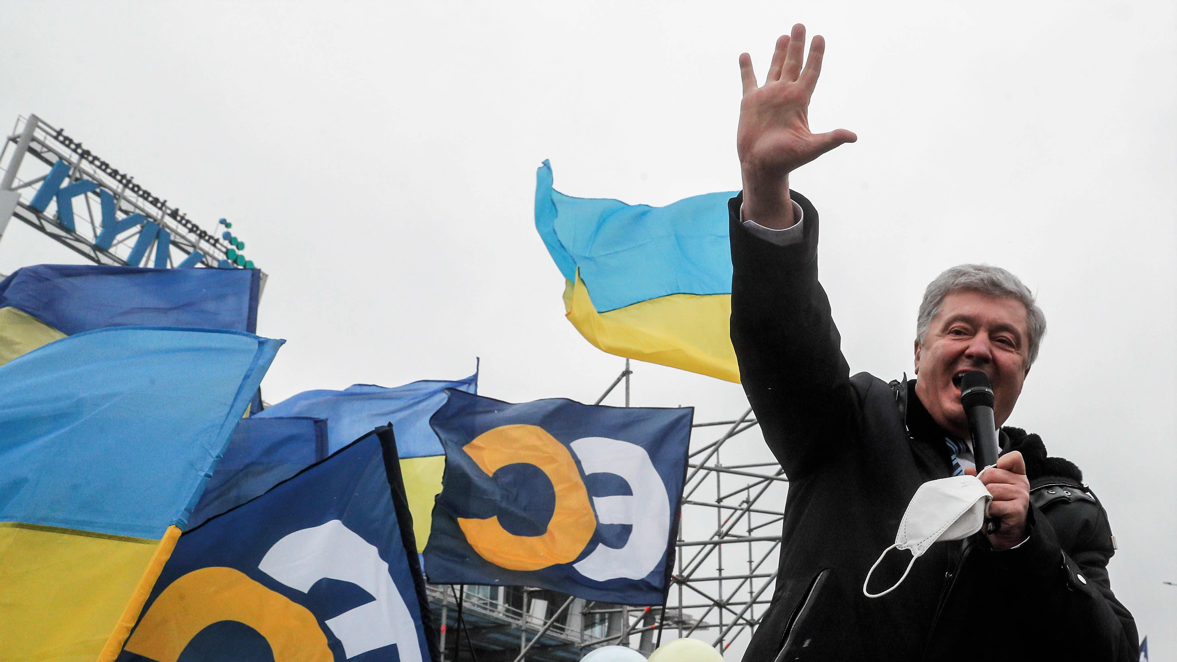 Poroshenko puhuu mikrofoniin ulkona. Hänellä on tumma päällystakki yllään. Poroshenko nostaa oikean kätensä korkealla ilmaan. Taustalla näkyy Ukrainan ja hänen puolueensa Eurooppalaisen solidaarisuuden lippuja sekä Kiovan lentokentän kyltti.