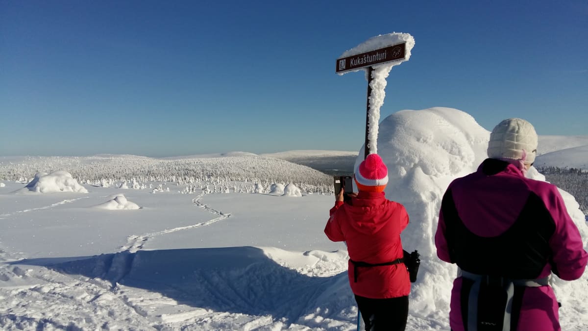  Kaksi punaiseen ja violettiin pukeutunutta hiihtäjää seisoo tunturin päällä Lapissa ja ottaa valokuvia.