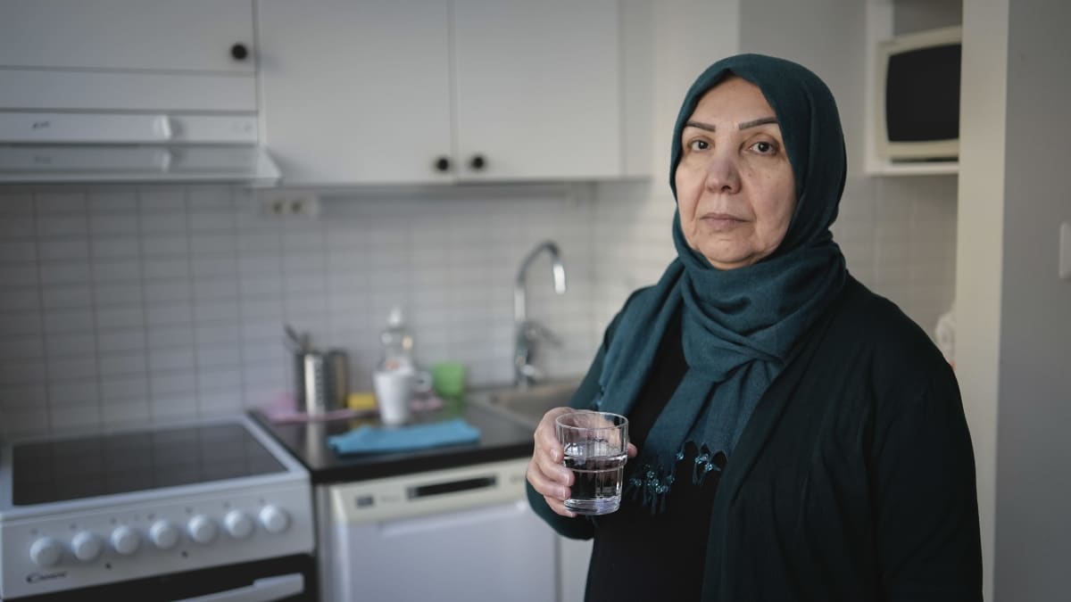 Huivipäinen nainen seisoo keittiössä vesilasi kädessään ja katsoo väsyneen näköisenä kameraan.
