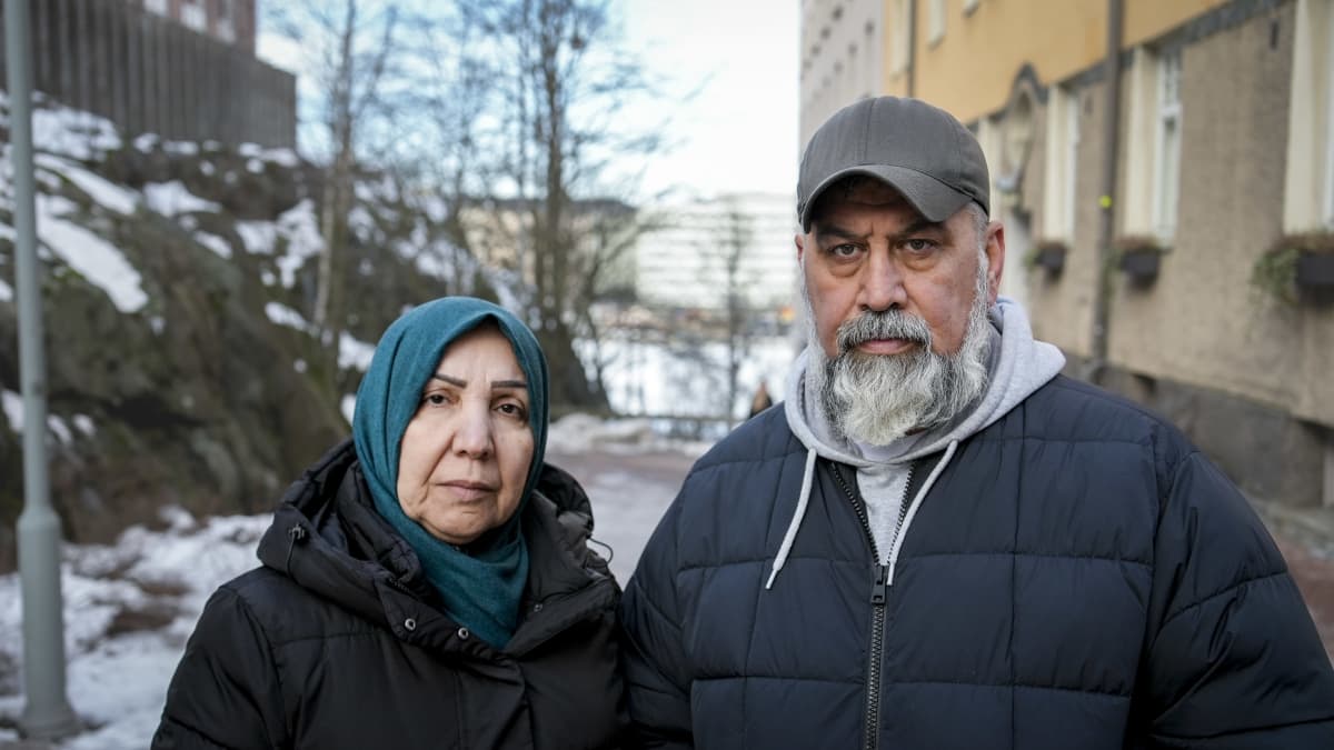 Keski-ikäinen, surullisenoloinen pariskunta seisoo ulkona ja katsoo kameraan.