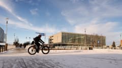 Paksurenkaisella pyörällä ajava polkypyöräilijä ajaa etualalla. Taustalla Oulun pääkirjasto.