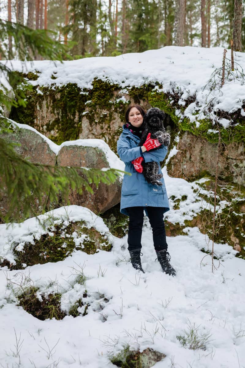 Tiina Raevaara pitää mustaa villakoiraa sylissään lumisessa metsässä kallioiden edessä.