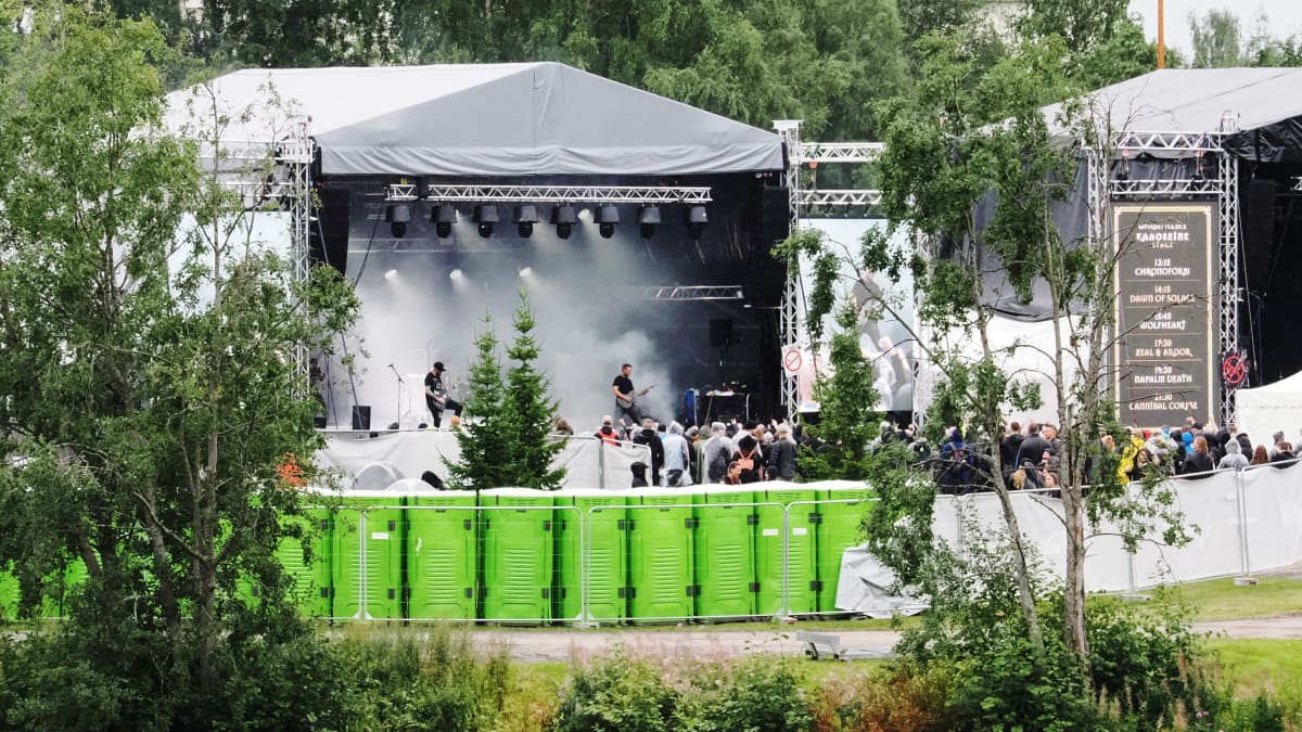 Rock bändi soittaa esiintymislavalla savutehosteiden keskellä ja edessä sadevarusteisiin pukeutunutta yleisöä. Etualalla rivissä vihreiden käymälöiden takaseiniä.
