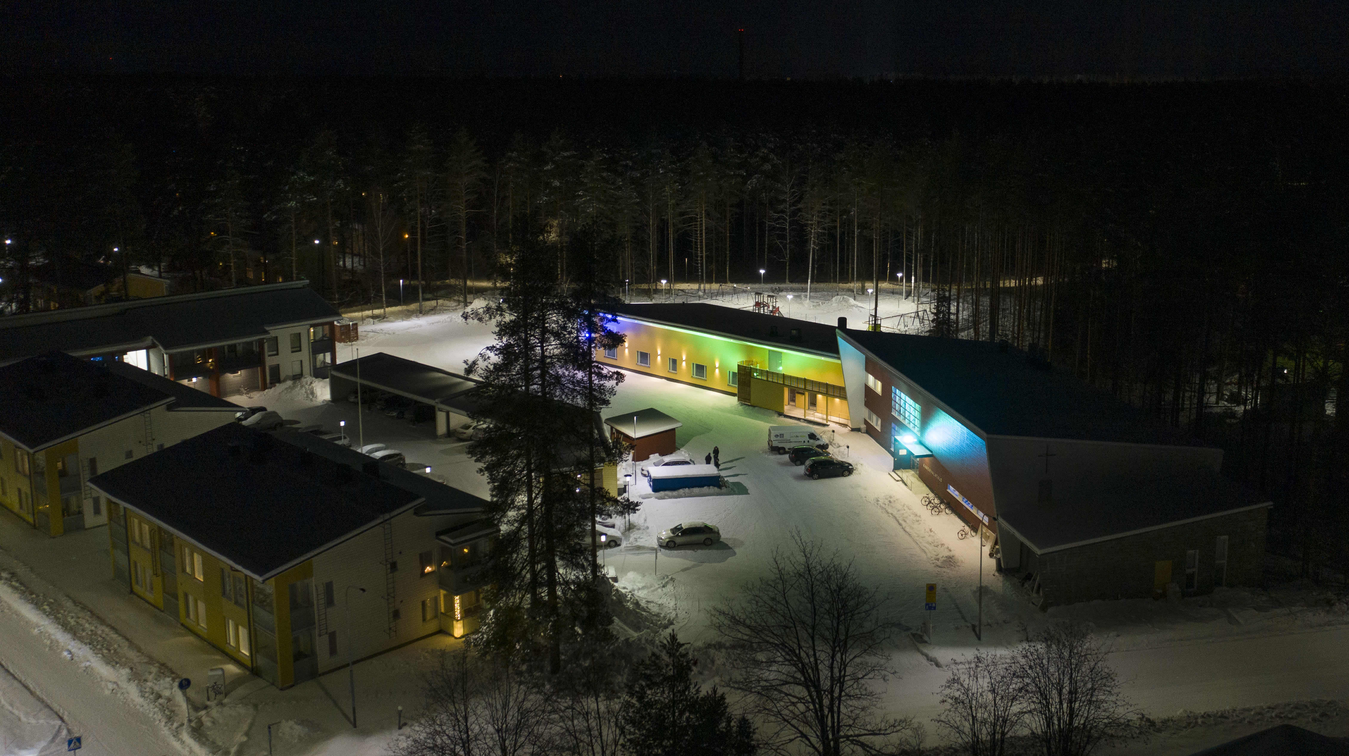 Joensuun Vapaaseurakunnan toimitilat loistavat värikkäissä koristevaloissa lumisessa Kanervalan kaupunginosassa. Ilmakuva, joka on otettu pimeällä.