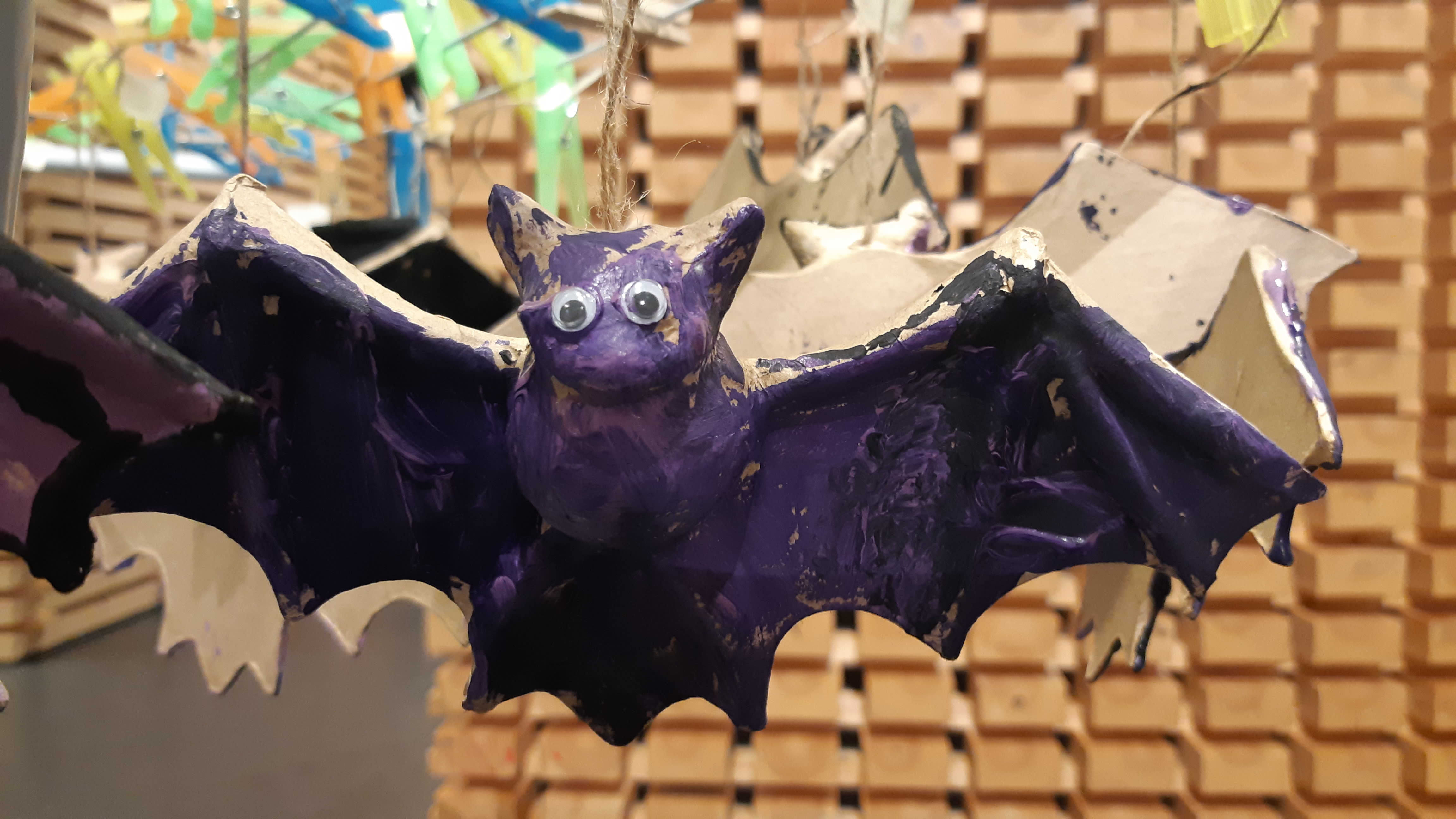 Lapsen askartelema ja violetiksi maalaama lentävä lepakko, jolla on muovisilmät. Halloween-koristeeksi tarkoitettuja maalattuja lepakoita on kuivumassa työpajassa pyykkipojilla pieneen telineeseen kiinnitettyinä.