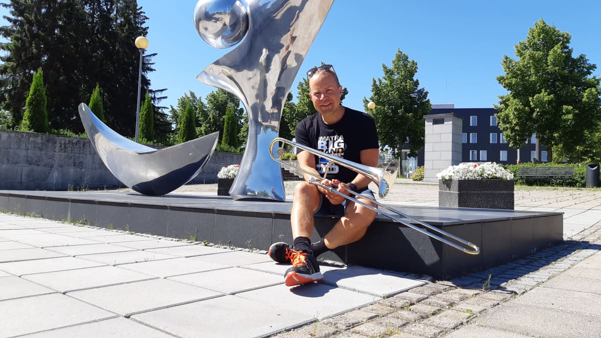 Muusikko Antti Rissanen istuu pasuunan kanssa metallisen patsaan edessä Imatran kulttuurikeskus Virran pihalla.