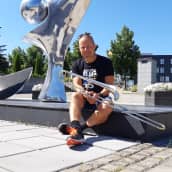 Muusikko Antti Rissanen istuu pasuunan kanssa metallisen patsaan edessä Imatran kulttuurikeskus Virran pihalla.