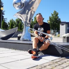 Muusikko Antti Rissanen istuu pasuunan kanssa metallisen patsaan edessä Imatran kulttuurikeskus Virran pihalla.