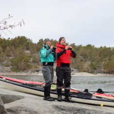 En kvinna och en man i friluftsutrustning står med kikare i handen på ett berg, två kanoter är uppdragna bakom dem. 