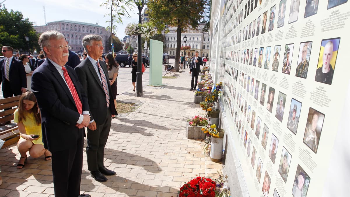 Mursuviiksinen John Bolton katselee toisen miehen kanssa muistoseinää, jossa on kaatuneiden ukrainalaisten kuvia. Kukkia seinän edessä. 