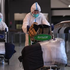 Kaksi suojapukuun pukeutunutta matkustajaa kävelee matkatavarakärryn kanssa Melbournen lentoasemalla. 