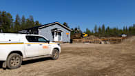 Small white truck on a gravel road, with a grey house, excavator and trees seen in the background.