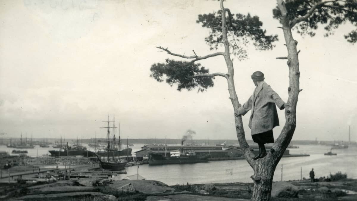 Nainen seisoo haarautuneen puun oksalla ja katsoo satamaa kohti. Satamassa muutamia laivoja ja rakennus.