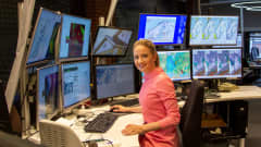 Meteorologi Anniina Valtonen hymyilee kameralle työpisteellään ja häntä ympäröi yhteensä 10 näyttöä, joilla on erilaisia sääkarttoja.