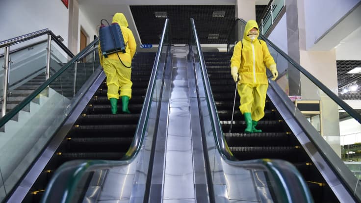 Kaksi suoja-asuista henkilöä vierekkäsissä liukuportaissa.