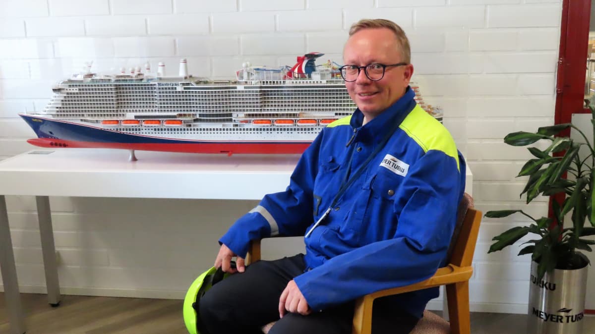 Mies istuu risteilijän pienoismallin edessä Meyerin Turun telakalla ja hymyilee.