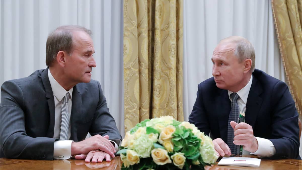 Viktor Medvedtšuk ja Putin istuvat pöydän ääressä, pöydällä on suuri keltavihreä kukka-asetelma. 