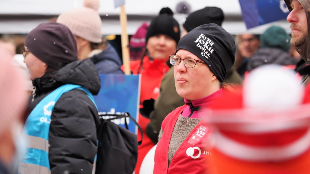 Punaiseen JHL:n liiviin pukeutunut nainen, jonka päässä on musta pipo, jossa teksti Kyllä joku hoitaa.  Taustalla mielenosoittajia.