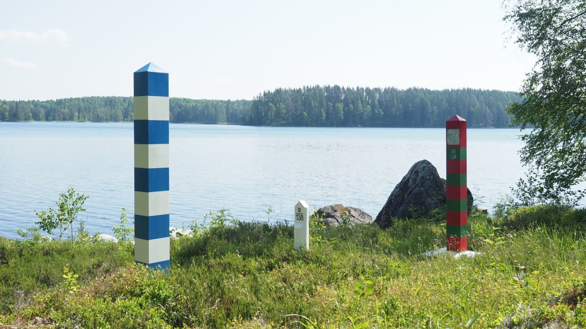 Suomen ja Venäjän rajamerkit vierekkäin maastossa järven rannalla.