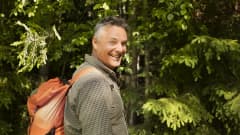 Mika Kalakoski seisoo hymyiillen metsän reunassa reppu selässään. 
